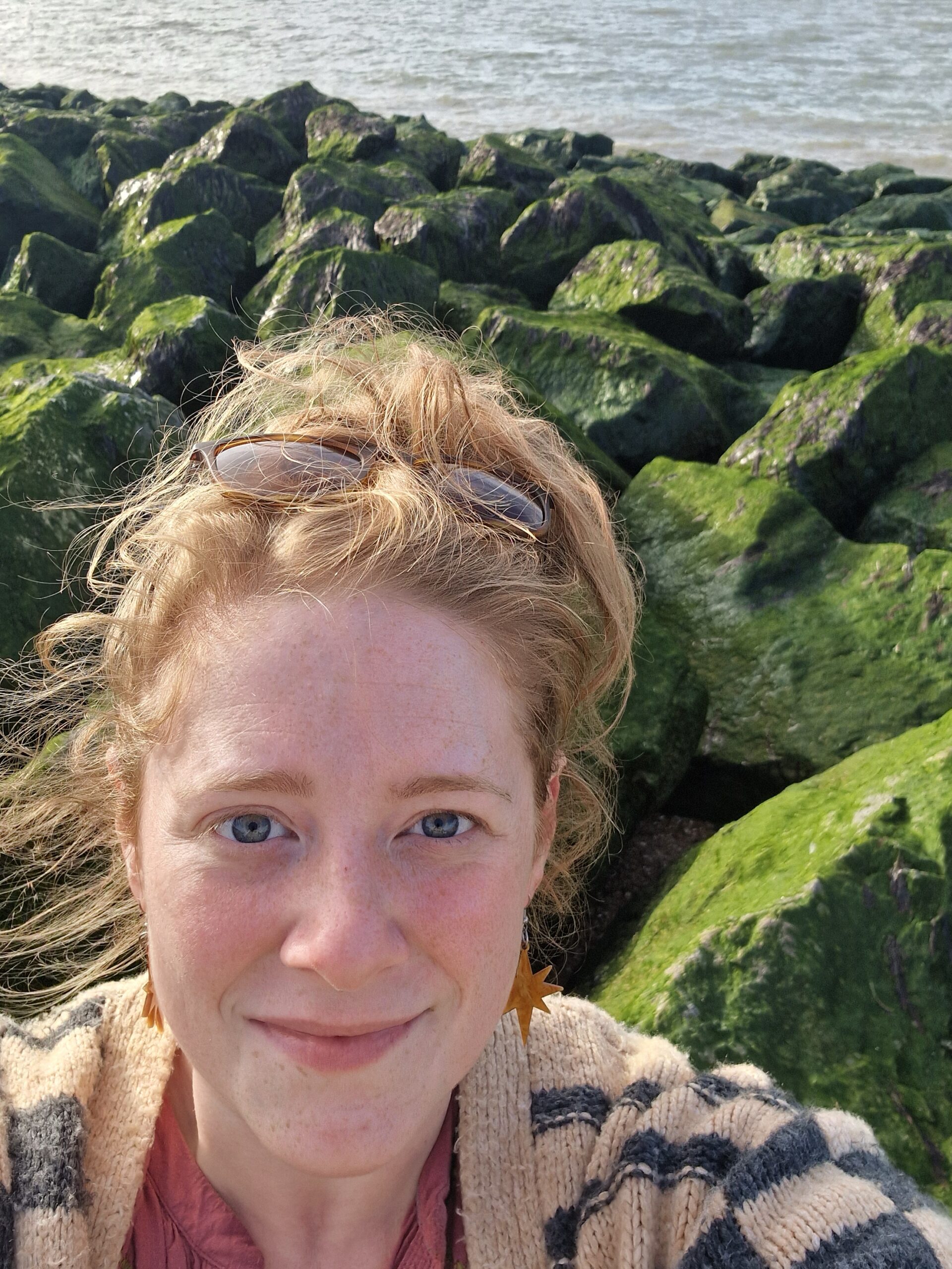 A white red headed woman smiles at the camera with sunglasses on her head, in the background are green algae covered rocks and the ocean.